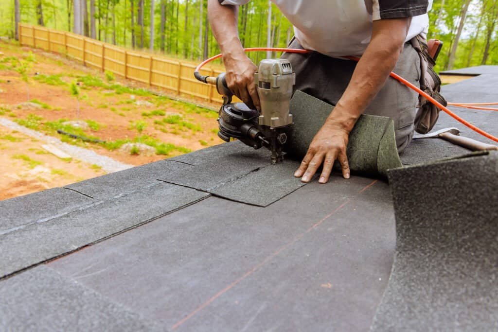 Man installing asphalt shingles on a rooftop with power tools, outdoor construction scene.